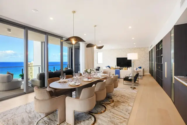 Open-plan dining and living with oval timber table, tan velvet chairs and sculptural black pendants over ocean views