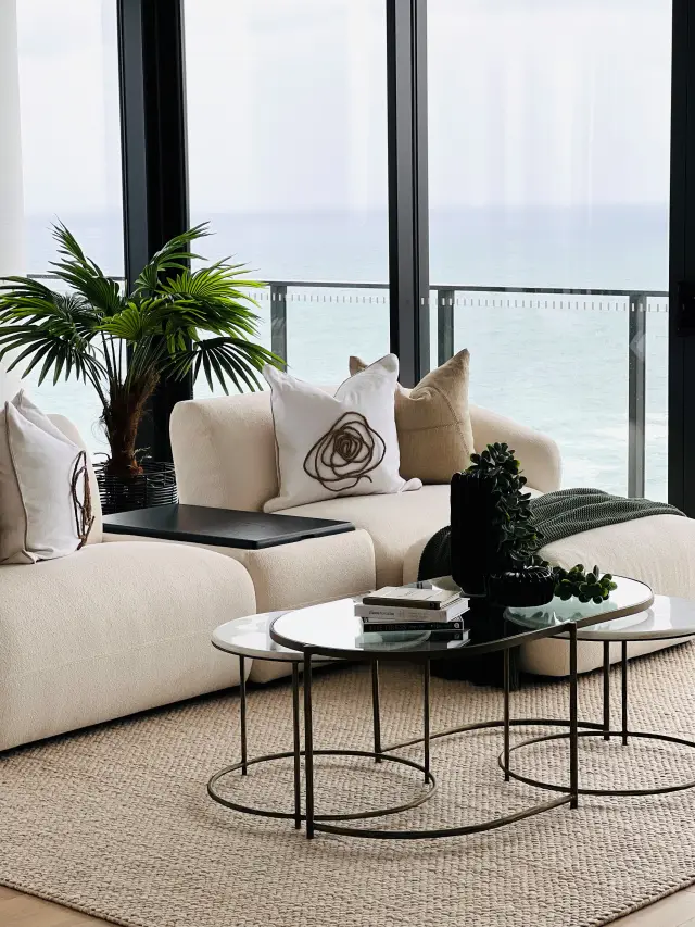 Living area detail with cream sofa, marble nesting tables and palm plant against ocean backdrop