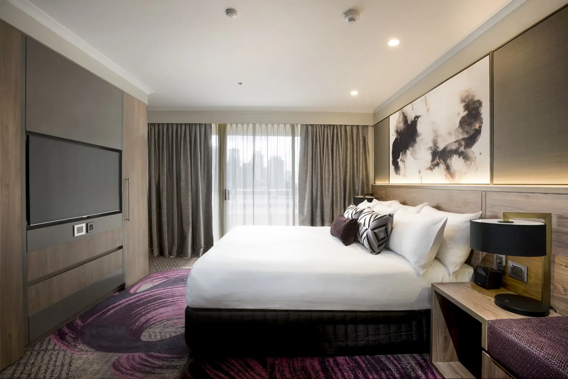 Guest suite at Rydges South Bank with a king bed, full-height timber joinery, patterned carpet and a city outlook through sheer curtains