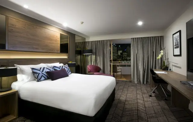 Deluxe king room with timber feature headboard, work desk and evening city skyline view