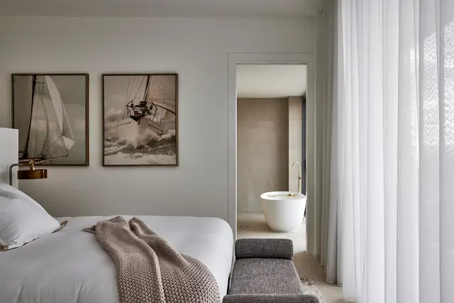 Bedroom with framed sailing artwork, knit throw and ensuite freestanding bath beyond sheer curtains