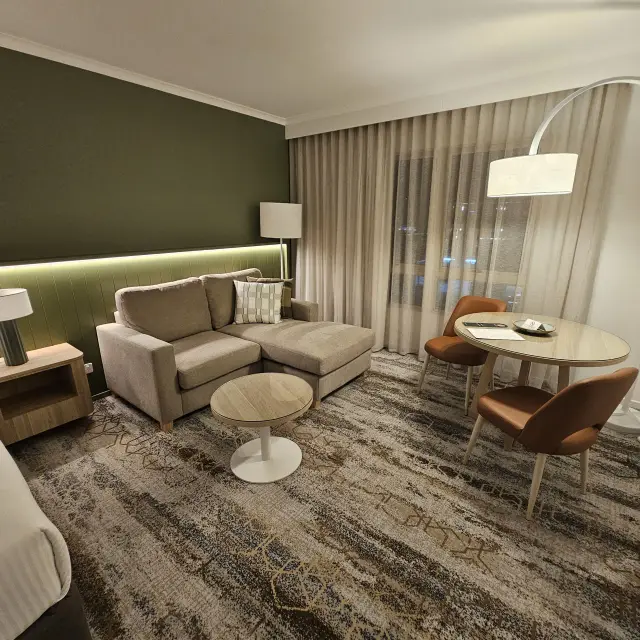 Guest room lounge zone furnished with a taupe chaise sofa, round coffee table, timber dining table and tan leather dining chairs