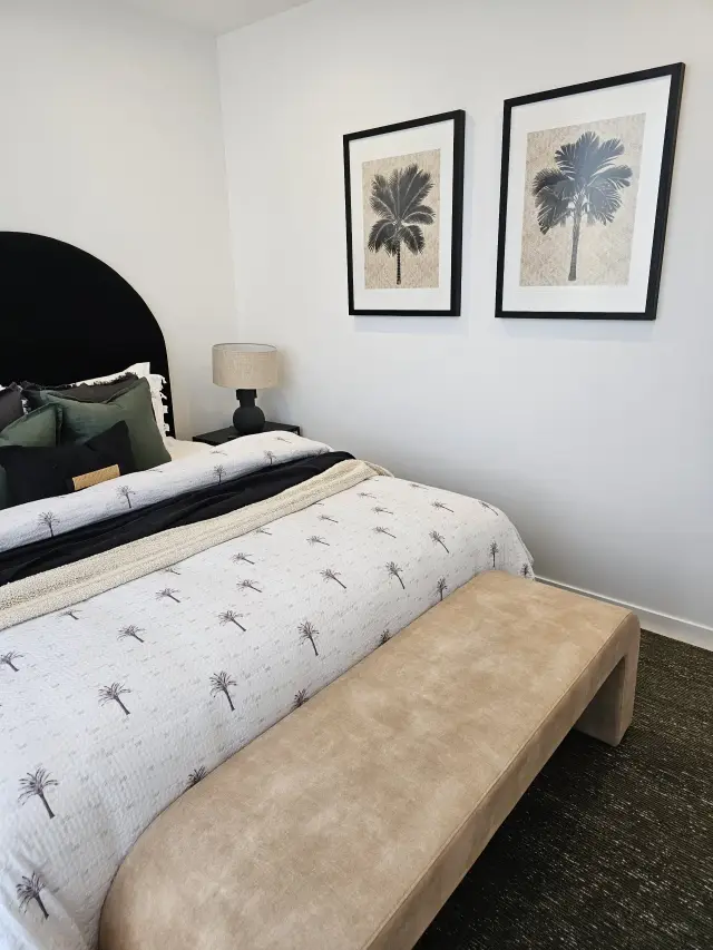 Bedroom with palm-print quilt, tan bench and framed palm-tree prints on white wall