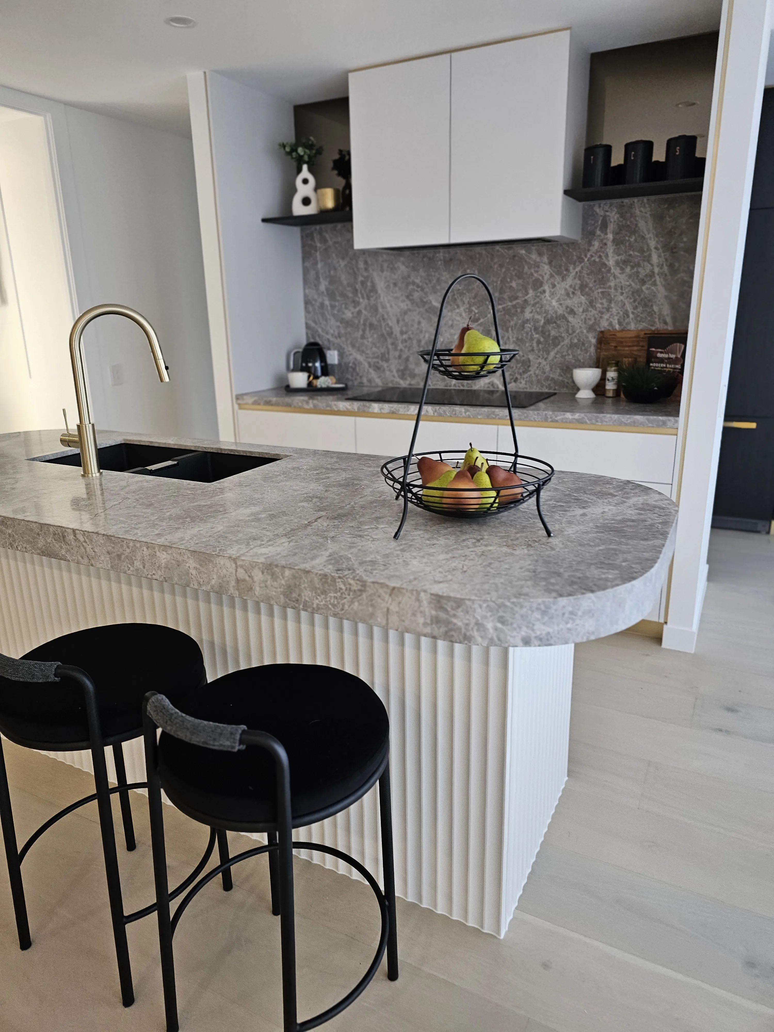 Kitchen with grey marble benchtop, fluted island panel, brass tapware and black bar stools
